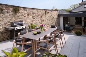 Une terrasse extérieure avec un mur en pierre, un barbecue, une table à manger avec des chaises et une vue sur la maison.