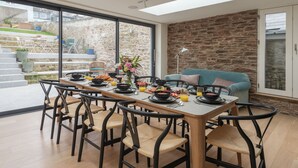 A modern dining area with a long table set for breakfast, surrounded by chairs, and a stone wall in the background.
