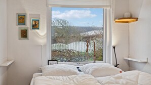A bedroom with a large window, white bedding, and a view of trees and a fence.