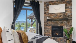Une chambre avec une cheminée en pierre, une grande fenêtre avec des rideaux et une vue sur l’extérieur.