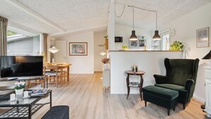 A modern living room with a flat-screen TV, a dining area with a wooden table and chairs, a green armchair, and a kitchen area with a microwave and plants. - Amazing home in Gørlev with WiFi (Gørlev)