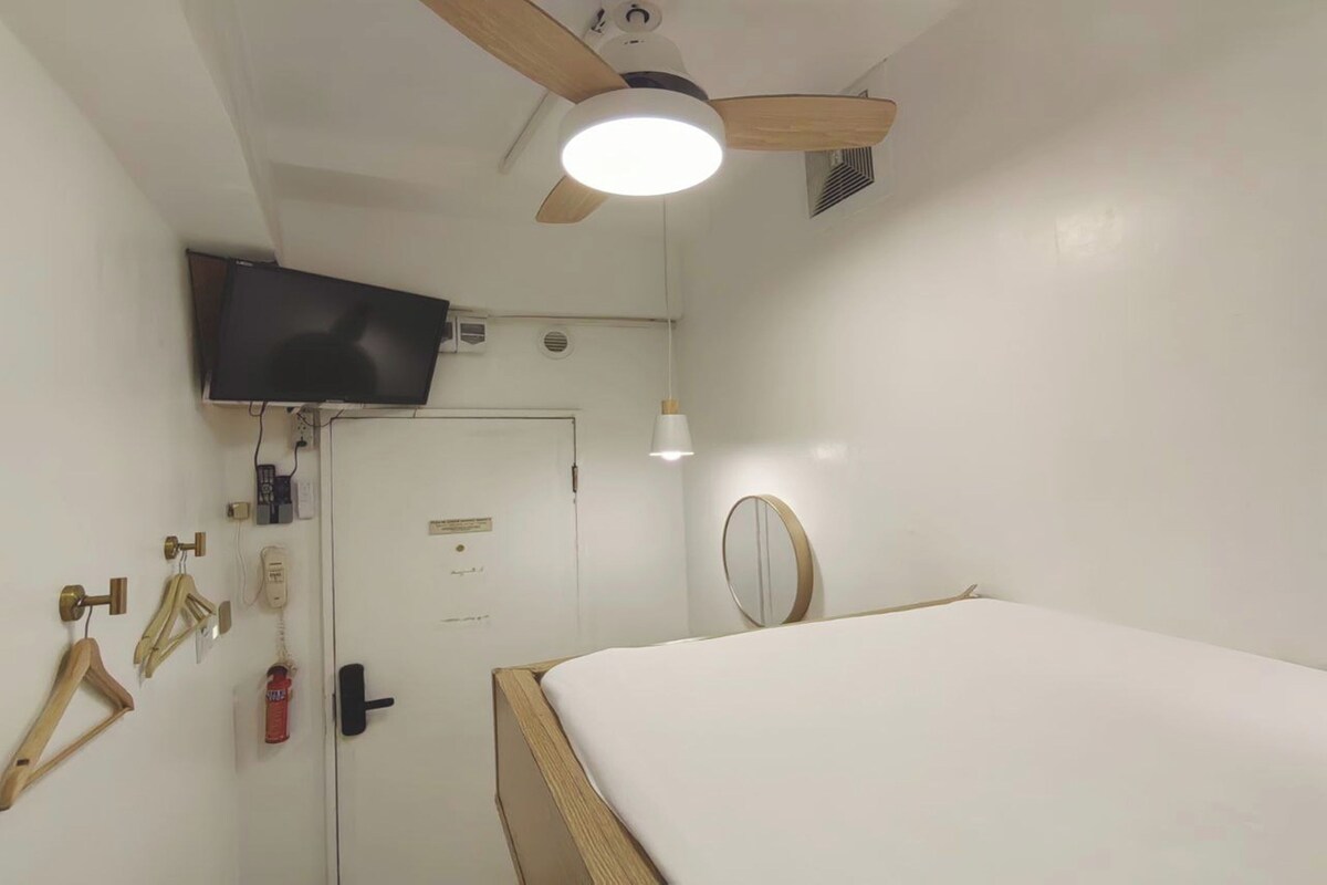 Habitación de hotel con cama, ventilador de techo, televisor montado, puerta con cerradura, interruptor de luz y un espejo pequeño.