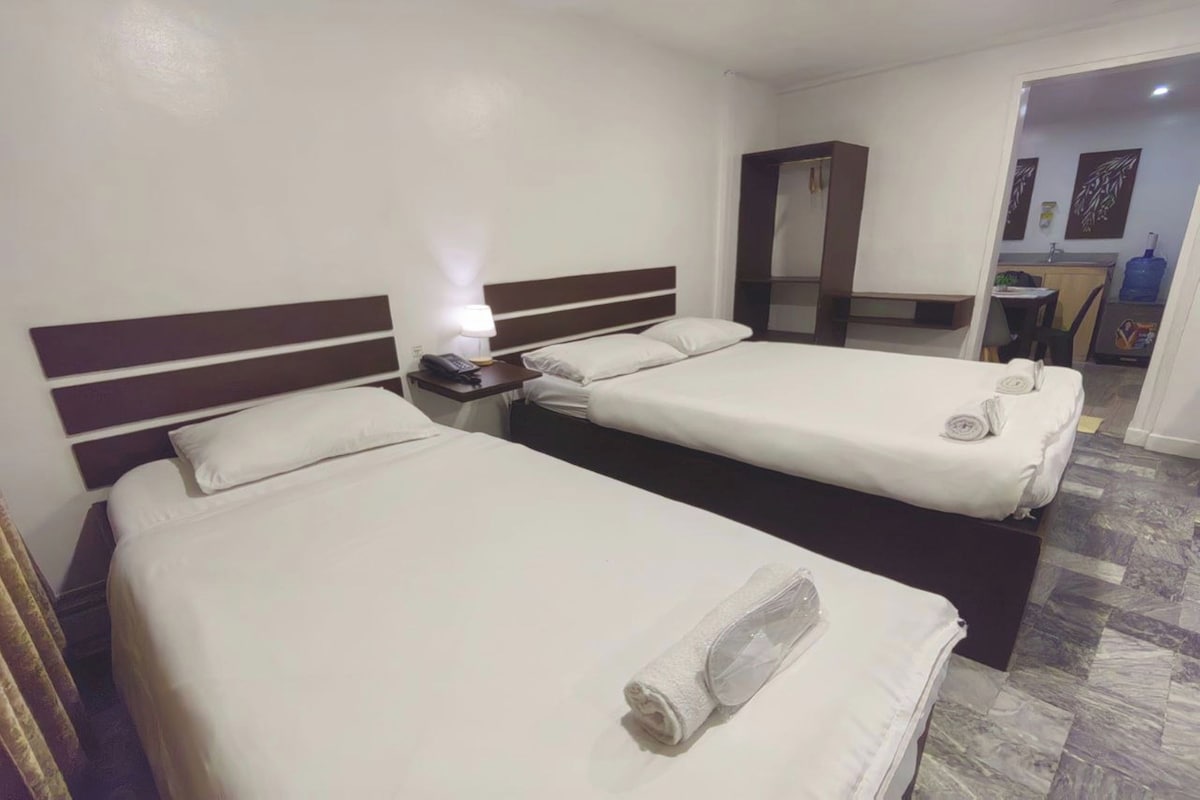 Habitación de hotel con dos camas individuales, una mesita de noche con lámpara, una pequeña mesa y un baño visible a través de una puerta abierta.