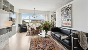 A modern living room with a black leather sofa, a coffee table, and a large window offering a view of buildings outside. - Gorgeous apartment in Fanø with WiFi (Fanø)