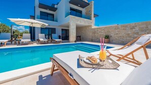 Uma casa moderna com dois andares, piscina, cadeiras de descanso e uma mesa com um croissant e bebida.