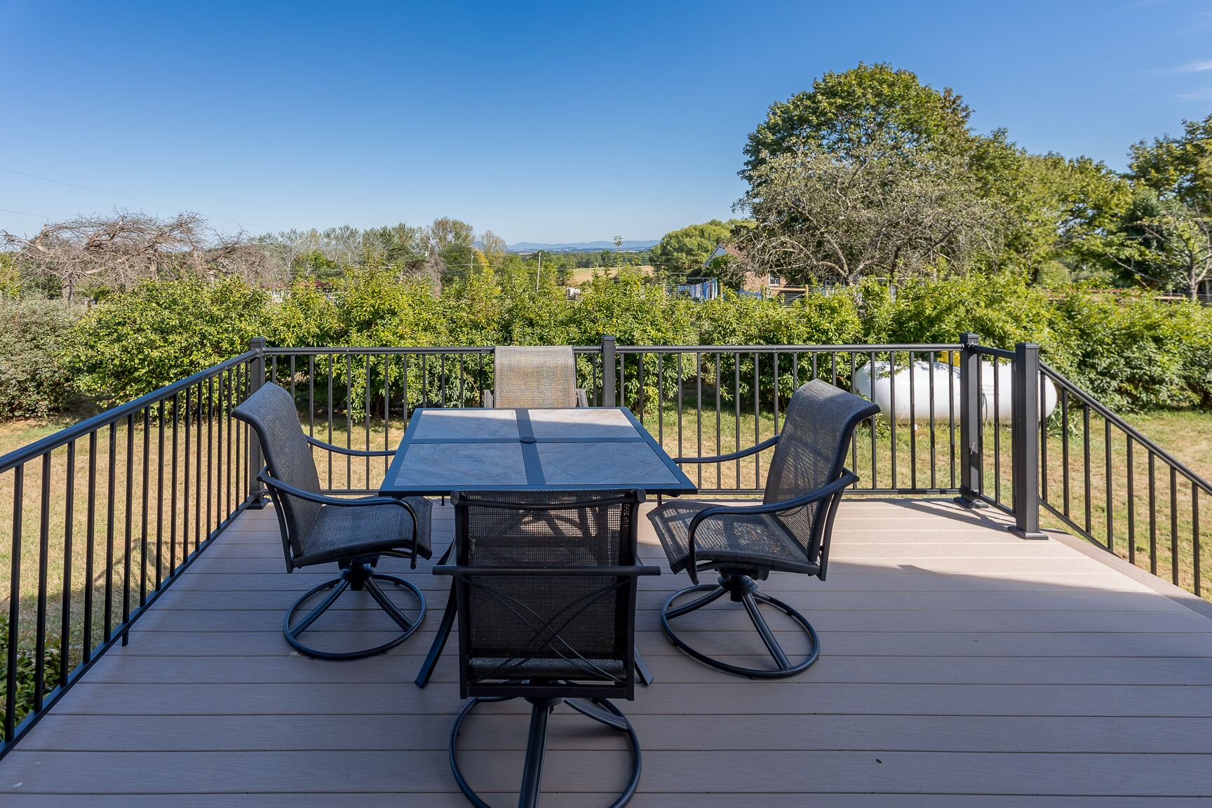 Eine Terrasse mit Tisch und vier Stühlen, umgeben von einem Metallgeländer, mit Blick auf einen angelegten Garten und weit entfernte Hügel.