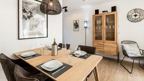 Uma área de refeições com uma mesa de madeira posta para quatro pessoas, cercada por cadeiras. Um armário com portas de vidro é visível ao fundo, e um relógio de parede está fixado na parede.