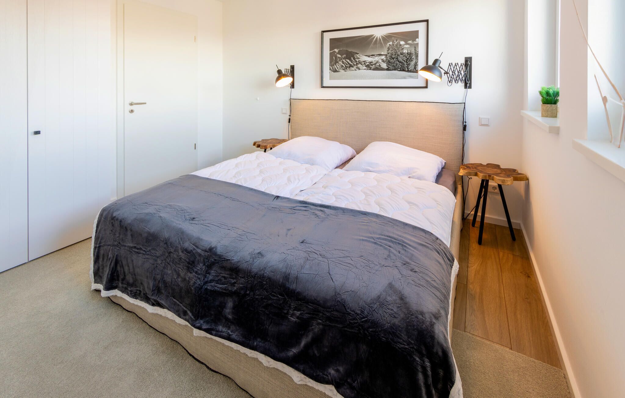 Ein Schlafzimmer mit Bett, Kopfstütze, Nachttisch, Lampe, gerahmtem Bild an der Wand und einer kleinen Pflanze auf einem Regal.