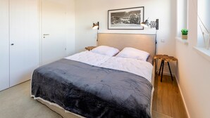 Quarto com cama, cabeceira, mesinha de cabeceira, abajur, quadro na parede e uma pequena planta em uma prateleira.