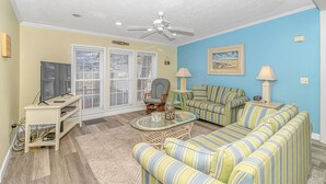 A living room with a TV, a ceiling fan, a glass coffee table, striped sofas, and a painting on the wall. - Large 3 bedroom in Ocean Lakes; sleeps up to 10, parking for up to 5 vehicles. (Myrtle Beach)