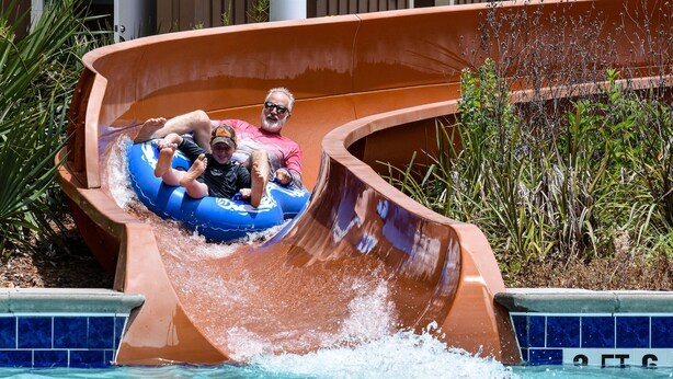 A water slide with a curved design, surrounded by a pool and greenery. - Large 3 bedroom in Ocean Lakes; sleeps up to 10, parking for up to 5 vehicles. (Myrtle Beach)