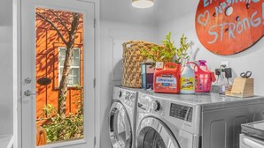 Een wasserette met een wasmachine, een droger, een brandblusser en een deur met uitzicht op een oranje gebouw.