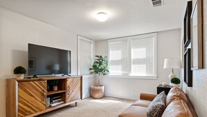 A living room with a flat-screen TV on a wooden cabinet, a brown leather sofa, a potted plant, and a window with blinds. - 2BR Downtown Sac - In unit Laundry - Free Parking (Sacramento)