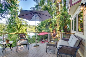 Une terrasse en bois avec des meubles en osier, un parasol rouge et une vue sur des arbres et un lac.
