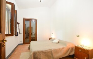 Un dormitorio con cama, mesita de noche, armario de madera, espejo y puerta que da a otra habitación.