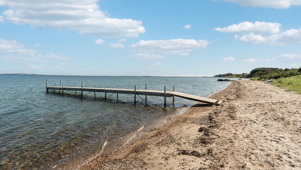 Un muelle de madera que se adentra en aguas tranquilas, una playa de arena y una costa lejana con vegetación.