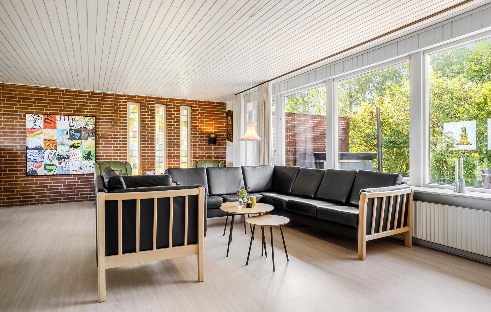 Un salon moderne avec un mur en briques, un canapé en cuir noir, deux fauteuils en bois, une table basse ronde et de grandes fenêtres donnant sur un paysage verdoyant.