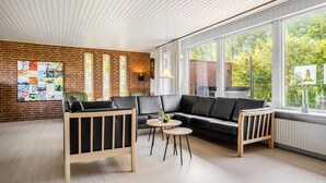Un salón moderno con pared de ladrillo, un sofá de cuero negro, dos sillones de madera, una mesa de centro redonda y grandes ventanales con vistas a zonas verdes.