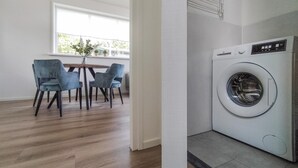 A laundry room with a front-loading washing machine, a dining area with a table and chairs, and a window with a view of greenery. - Lovely home in Oosterwolde with WiFi (Oosterwolde)