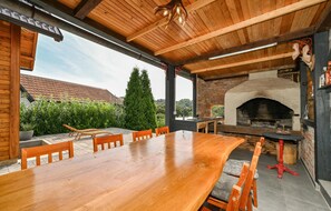 A wooden dining table with chairs, a brick oven, and a view of a house with a tiled roof.