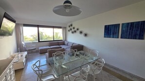 A modern living room with a glass dining table, white chairs, a sofa, and a large window with curtains. - Breathtaking 2 bedroom Condo at Ocean Front Property Walk To La Isla Mall (CancÃºn)