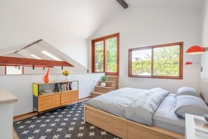 A bedroom with a bed, a nightstand, a bookshelf, and a window with a view of greenery.
