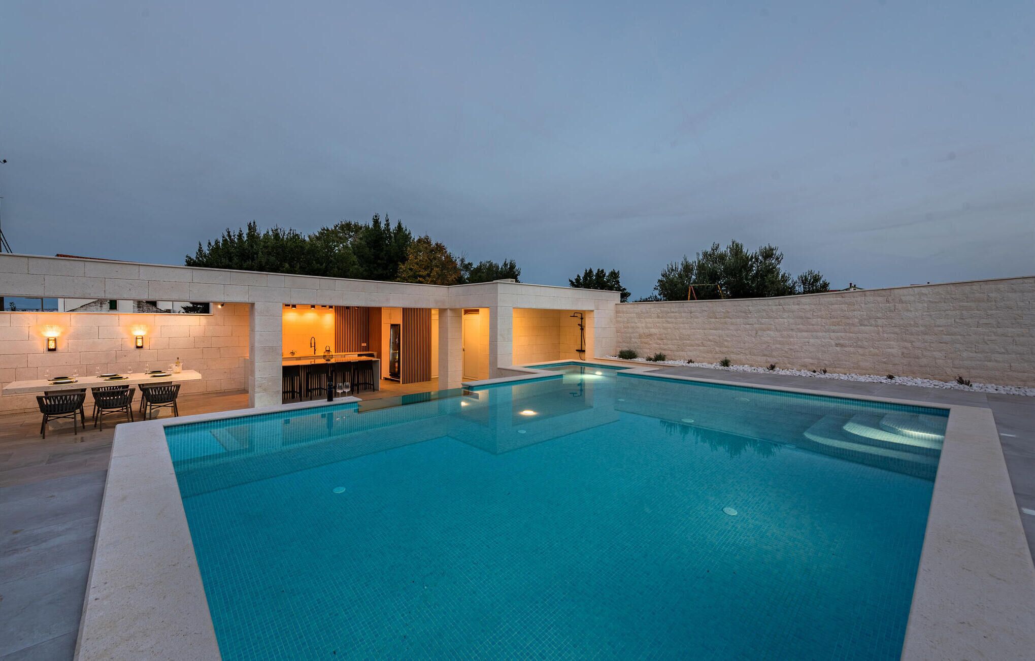A modern outdoor pool area with a bar and seating, surrounded by a stone wall and trees.
