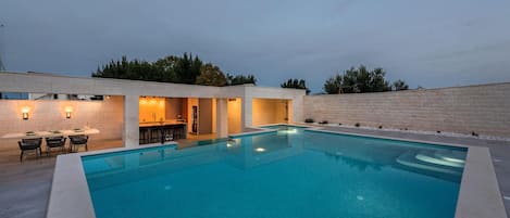 A modern outdoor pool area with a bar and seating, surrounded by a stone wall and trees.