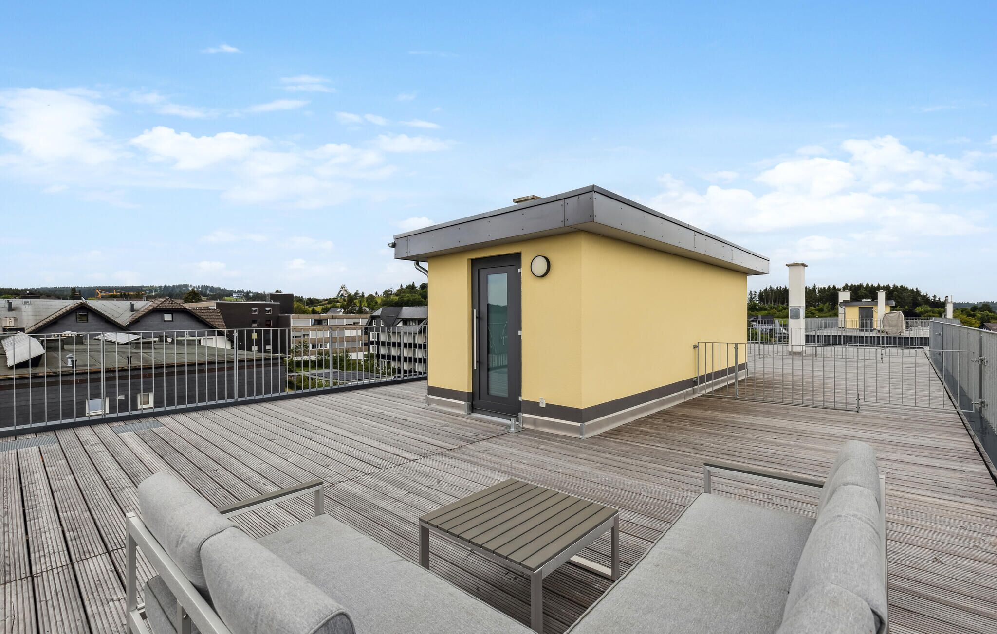 Une terrasse sur le toit avec un petit bâtiment jaune, une plateforme en bois et une rambarde en métal.