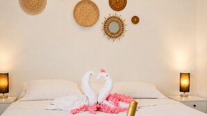 Ein Bett mit weißen Leinen, zwei Nachttischlampen, eine Champagnerflasche in einem Metallkübel und zwei Gläser auf einer gewebten Unterlage.