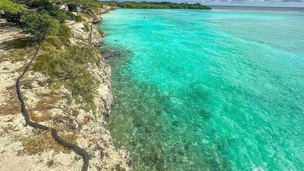 Ein Strand mit klarem türkisblauem Wasser, einem sandigen Ufer und einer entfernten Insel.