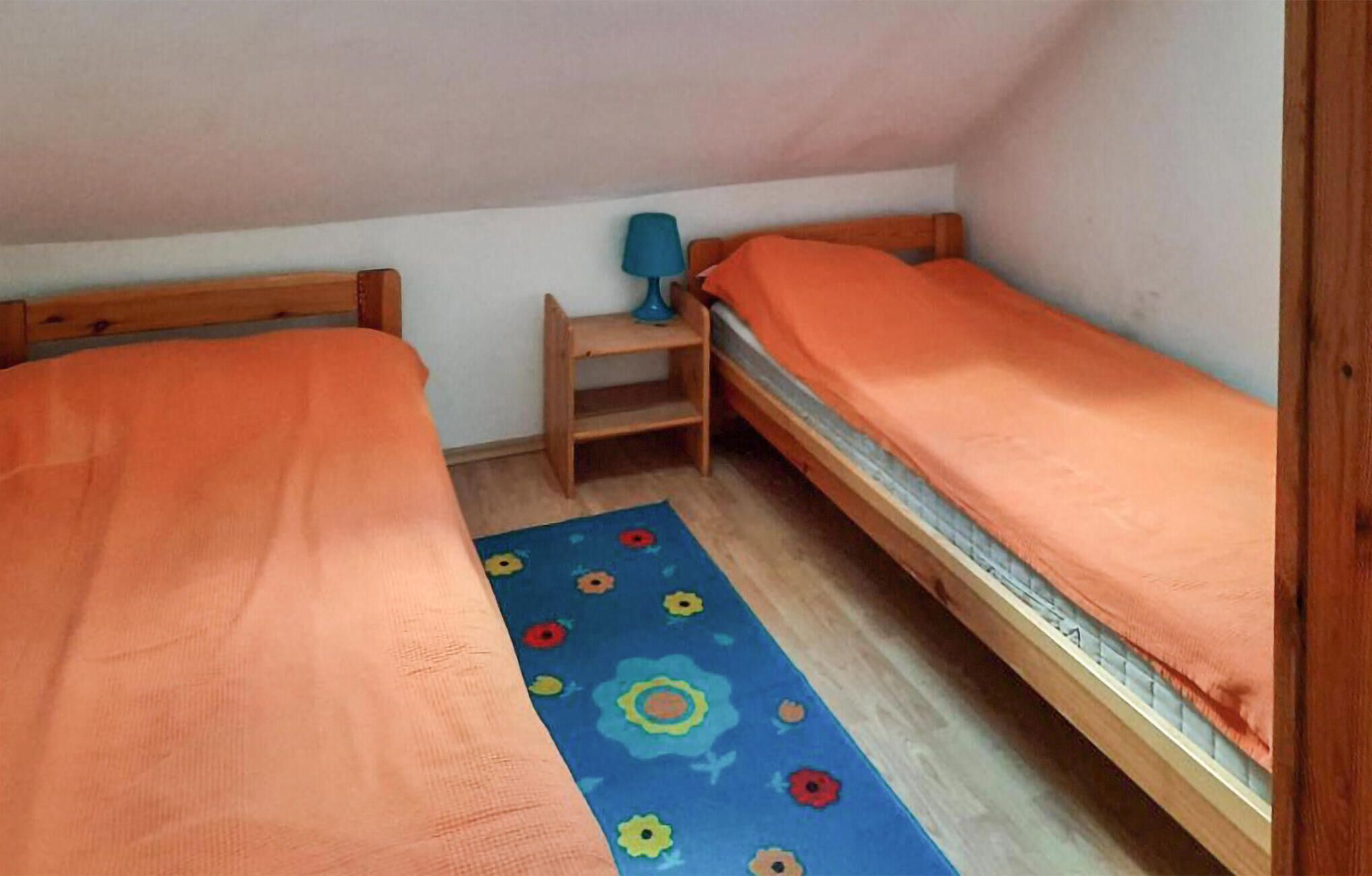 A small, single-room accommodation with two wooden beds, a blue floral rug, and a small table with a lamp.
