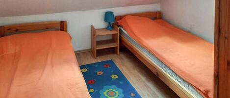 A small, single-room accommodation with two wooden beds, a blue floral rug, and a small table with a lamp.