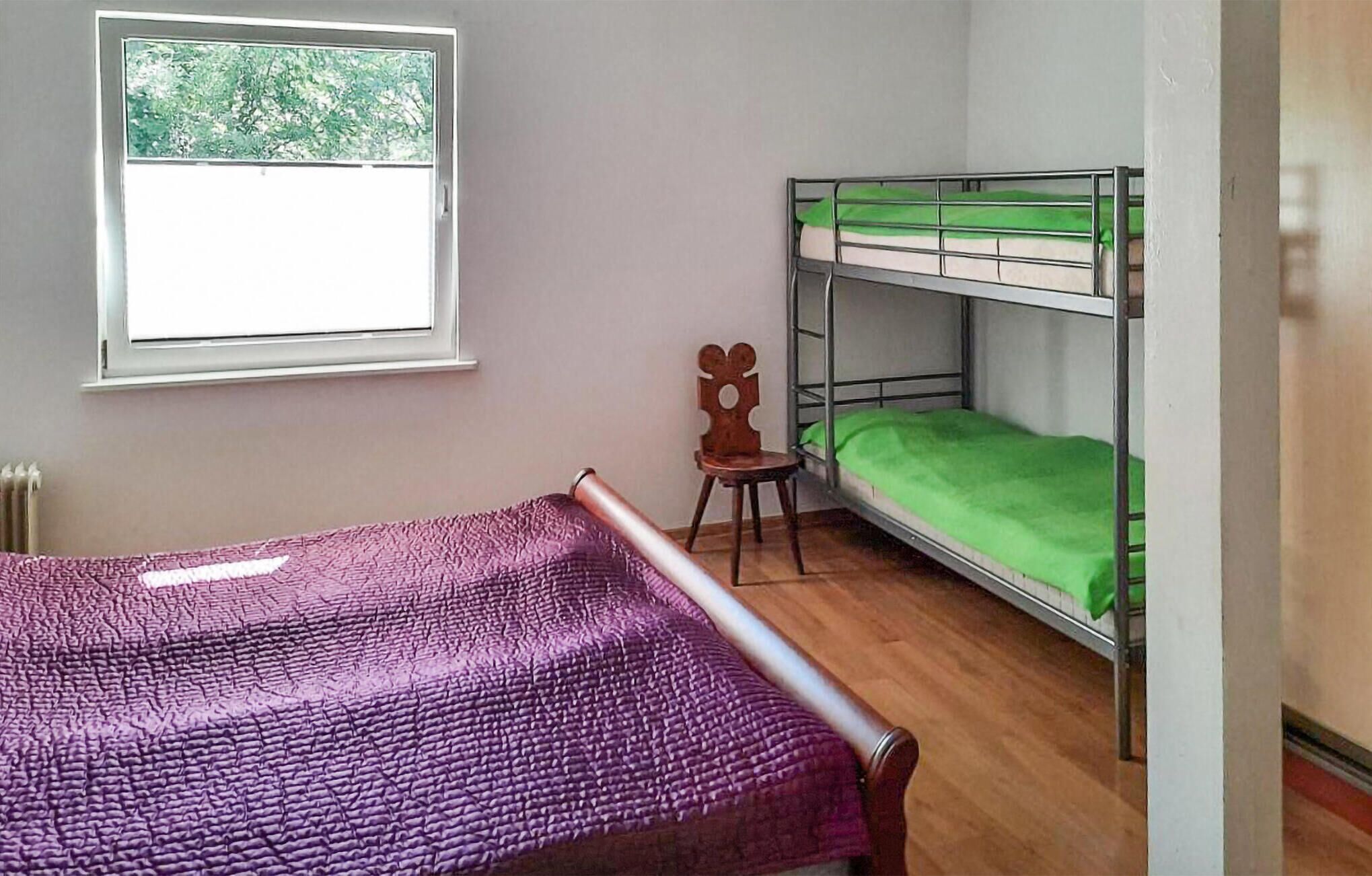 A room with a bunk bed, a single bed, a chair, a window with a view of trees, and a radiator.