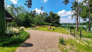 Un sendero pavimentado que lleva a una terraza de madera con un columpio, rodeado de abundante follaje y árboles altos, con vista a un lago.