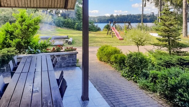 Una terraza de madera con un banco de piedra y una mesa de picnic, rodeada de exuberante vegetación y vistas a un lago con un columpio.