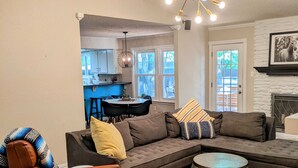 A modern living room with a sectional sofa, a leather armchair, and a round coffee table. There is a dining area visible through an open doorway, a fireplace, and a ceiling light fixture. - South Austin Charmer with Backyard Oasis (Austin)