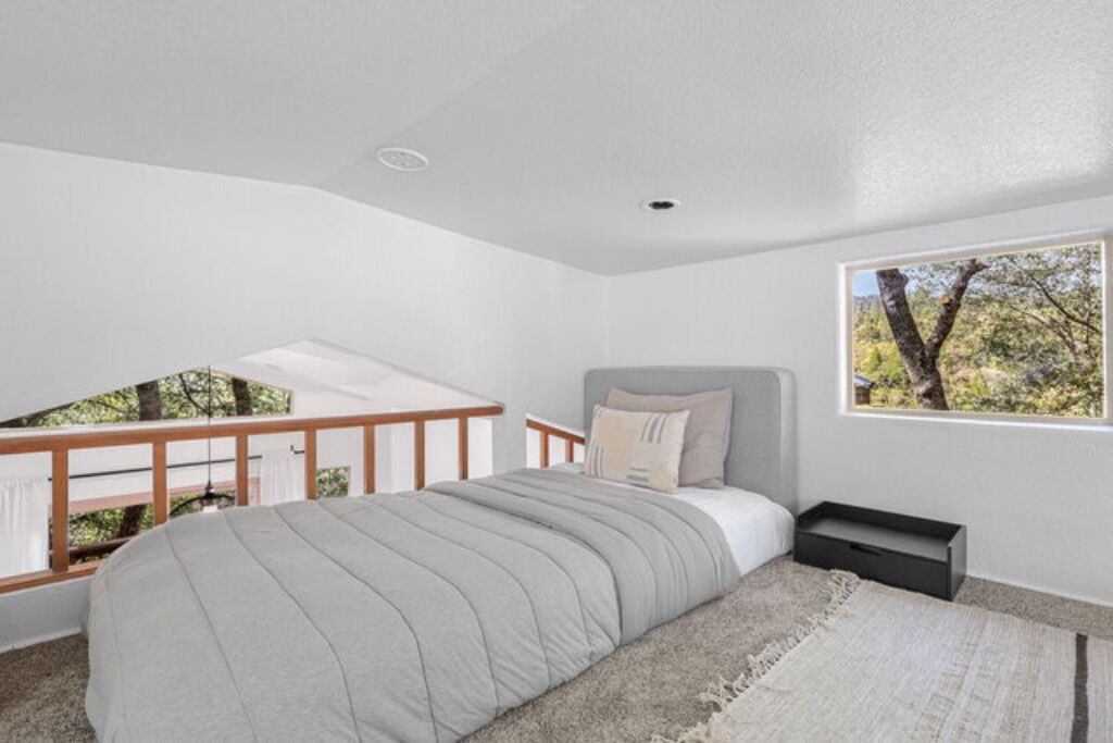 Quarto com cama, mesa de cabeceira, janela com vista para árvores e varanda com guarda-corpo.