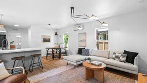 A modern living room with a sofa, coffee table, and bar stools. There is a kitchen area with a counter and stools. - Modern Cabin (Alder) (Dunsmuir)