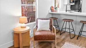 A modern kitchen with white cabinets, a wooden countertop, and a hanging pendant light. There is a brown leather chair with a white pillow, a round wooden side table with a lamp, and two bar stools. - Modern Cabin (Alder) (Dunsmuir)
