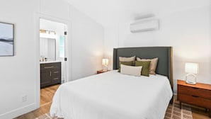 A bedroom with a large bed, bedside tables, a nightstand, a mirror, and an air conditioning unit. - Modern Cabin (Blue Spruce) (Dunsmuir)