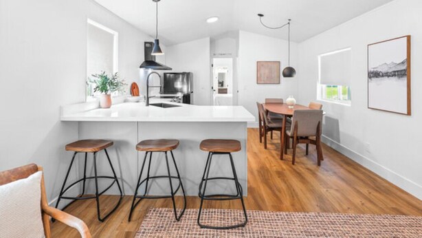 A modern kitchen with a white countertop, bar stools, and a dining area with a table and chairs. - Modern Cabin (Blue Spruce) (Dunsmuir)