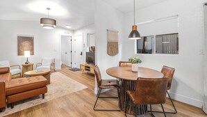 Un salon moderne avec une table à manger ronde en bois, des fauteuils en cuir et un canapé. Une grande fenêtre est agrémentée de rideaux, un luminaire suspendu éclaire la pièce et un tapis recouvre le plancher en bois.