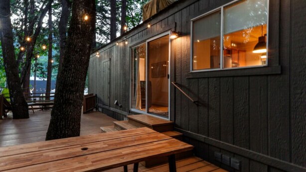 Une terrasse en bois avec une table et des bancs, entourée d’arbres, et un bâtiment doté d’une porte-fenêtre et de fenêtres coulissantes.