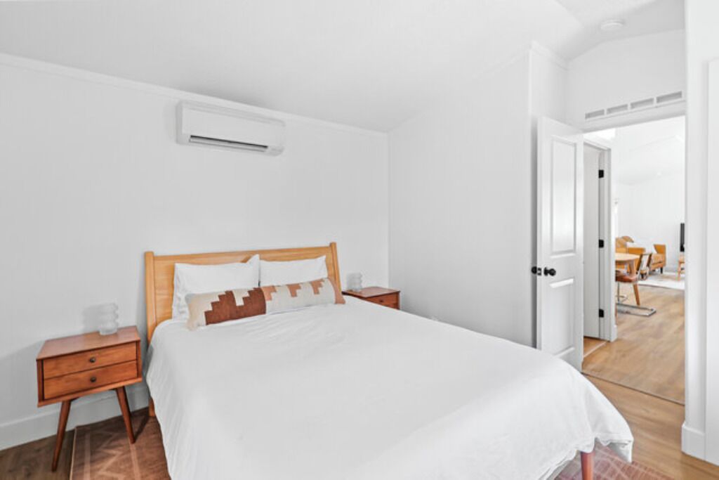 A bedroom with a wooden bed, white bedding, a nightstand, a drawer, and an air conditioning unit on the wall.