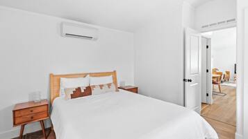 A bedroom with a wooden bed, white bedding, a nightstand, a drawer, and an air conditioning unit on the wall.