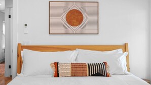 Een net opgemaakt bed met een kussen met patroon, een houten hoofdbord en een ingelijst kunstwerk erboven.