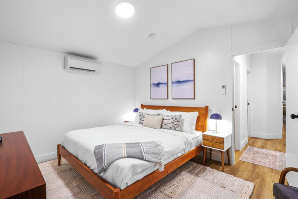 Une chambre à coucher avec un lit en bois, des murs blancs, deux tableaux encadrés, une table de chevet avec une lampe et un petit bureau en bois.
