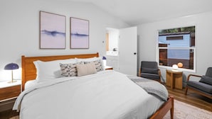 Una camera da letto con un letto grande, due quadri incorniciati, una scrivania con una lampada e una finestra vista su un edificio blu.