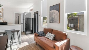 A modern kitchen with a stainless steel refrigerator, a dining area with a brown leather sofa, and a large window providing a view of the outdoors. - Tiny Home  - (Beech) (Dunsmuir)